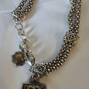 May include: Silver-toned bracelet with a double strand beaded design. The bracelet features decorative floral accents and two square pendants with ornate gold-colored filigree patterns. A small heart charm is also present.