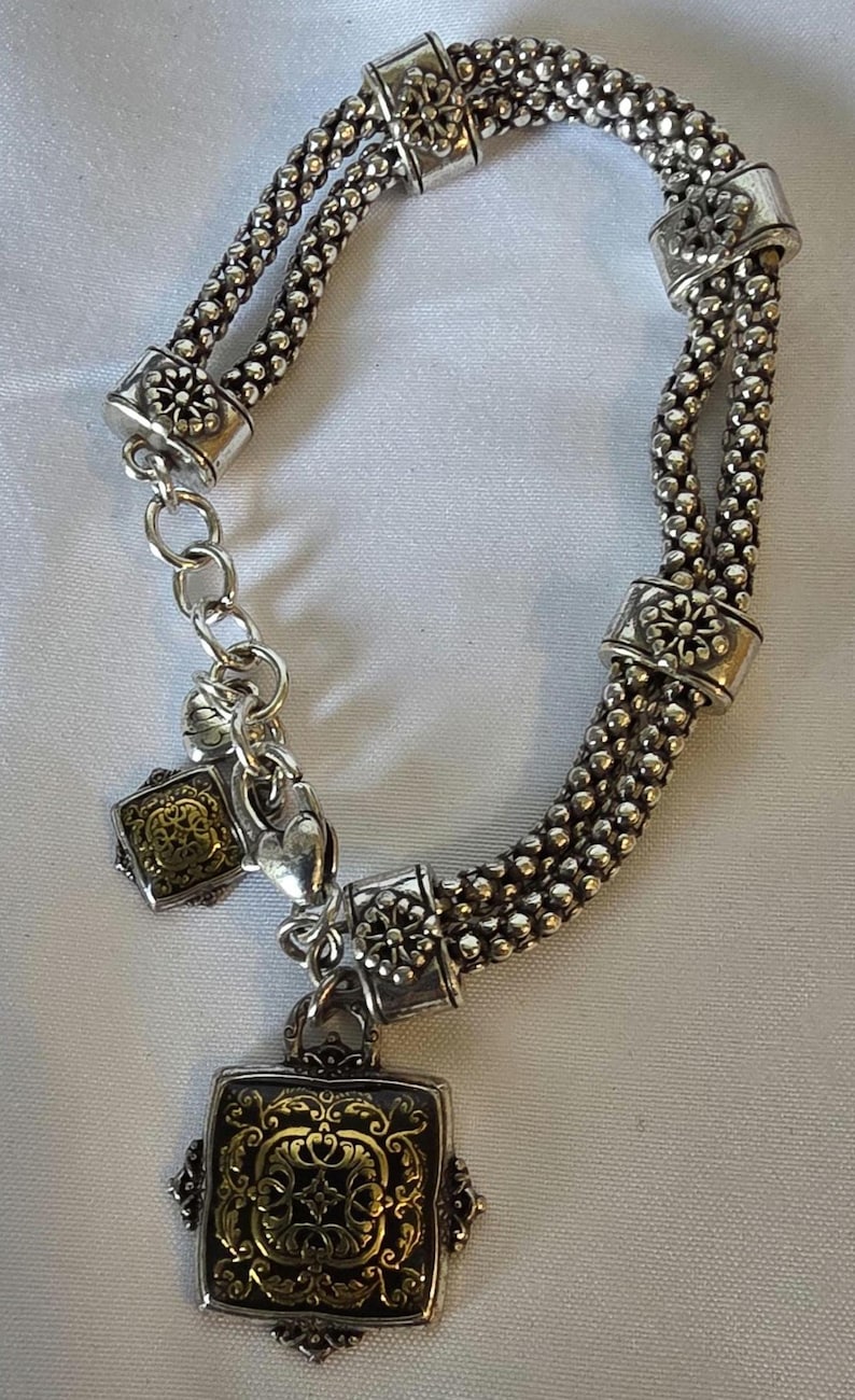 May include: Silver-toned bracelet with a double strand beaded design. The bracelet features decorative floral accents and two square pendants with ornate gold-colored filigree patterns. A small heart charm is also present.