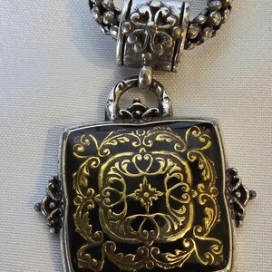 May include: Silver-toned necklace featuring a square pendant with a black background and ornate gold filigree design. The pendant is framed in silver and has decorative corner accents. The chain is a textured, silver-toned rope.