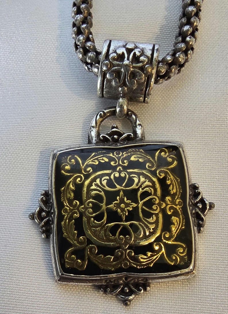 May include: Silver-toned necklace featuring a square pendant with a black background and ornate gold filigree design. The pendant is framed in silver and has decorative corner accents. The chain is a textured, silver-toned rope.