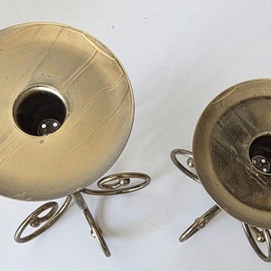 May include: Two vintage brass candle holders. Each has a circular base with a central opening for a candle, supported by delicate, curved metal legs. The brass has a warm, aged patina, adding to their antique charm.