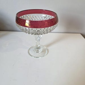 May include: A clear glass dessert bowl with a ruby red rim. The bowl has a diamond-cut pattern and sits on a clear stem and base. The bowl is likely used for serving desserts or small portions.