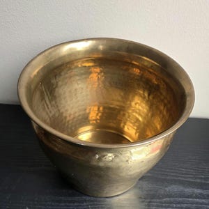 May include: A round, deep bowl-shaped planter crafted from polished brass with a distinct hammered texture across its entire surface. The vessel features a wide, smooth rim and a tapered base, showcasing a warm, metallic gold-toned finish.