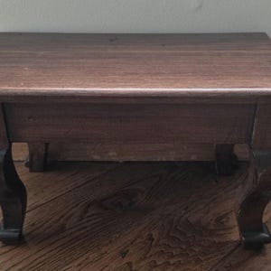 May include: A small, rectangular wooden stool featuring a smooth, dark brown top surface and four curved, cabriole-style legs. The stool is constructed from wood with a visible grain pattern, showcasing a consistent dark brown finish throughout the piece.