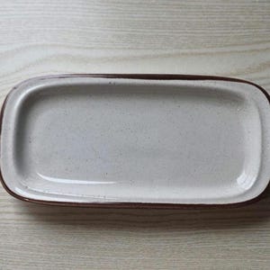 May include: Rectangular ceramic serving dish with a speckled off-white interior and a dark brown rim. The dish has rounded corners and a slightly raised edge. The neutral color palette makes it suitable for various food presentations.