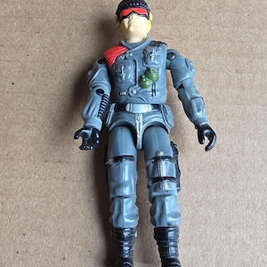 May include: A gray action figure of a soldier wearing a black helmet with red accents and yellow hair. The figure has a red shoulder pad, a green grenade, and black boots. The figure is wearing a gray uniform with black gloves.