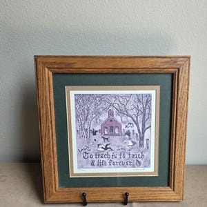 May include: A framed print featuring a schoolhouse illustration with trees, children, and a dog. The text reads, To teach is to touch a life forever. The artwork is set within a dark green mat and a light brown inner border, enclosed in a medium-toned oak wood frame.