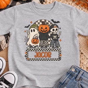 May include: A heather gray t-shirt featuring a graphic of a ghost, a pumpkin-headed figure, and a skeleton, all wearing hats and sneakers. The design includes bats, a spiderweb, and a rectangular box with the name JACOB in orange letters.