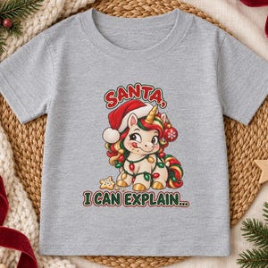 May include: A heather gray short-sleeve t-shirt featuring a graphic of a cartoon unicorn wearing a Santa hat, wrapped in colorful Christmas lights, and holding a red ornament. The text reads SANTA, I CAN EXPLAIN... above and below the unicorn.