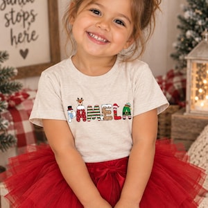 May include: A light heather gray short-sleeve t-shirt featuring the name PAMELA printed in a festive Christmas-themed font. Each letter is decorated with holiday motifs including a snowman, reindeer, candy stripes, and an elf hat.