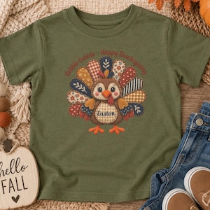 May include: An olive green short-sleeve t-shirt featuring a central graphic of a turkey with patchwork-style feathers in various patterns and colors. The text "Gobble Gobble" and "Happy Thanksgiving" arches above, with the name "Easton" on the turkey.