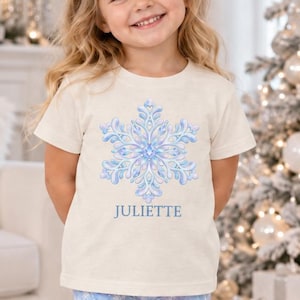 May include: A cream-colored short-sleeve t-shirt featuring a blue and white snowflake graphic with the name JULIETTE printed below it, paired with light blue leggings decorated with a white snowflake pattern and beige faux-fur lined boots.