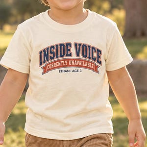 May include: A cream-colored short-sleeve t-shirt featuring the text INSIDE VOICE in dark blue with a red outline, followed by the text CURRENTLY UNAVAILABLE on a red banner, and the text ETHAN AGE 3 in dark blue lettering centered below.