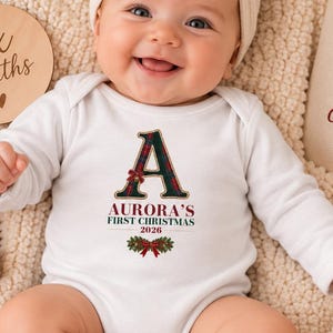 May include: A white long-sleeve baby bodysuit featuring a plaid letter A, the text Aurora's First Christmas 2026, and a holly graphic. The baby wears a cream-colored bow headband and red socks with bows, surrounded by a wooden six months sign.