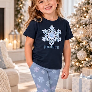 May include: A navy blue short-sleeved t-shirt featuring a large white and light blue snowflake graphic with the name JULIETTE printed below it, paired with light blue leggings covered in a repeating pattern of white and light blue snowflakes.