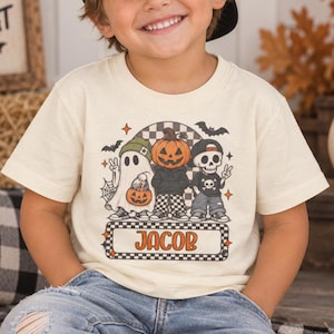 May include: A cream-colored short-sleeve t-shirt featuring a graphic print of a ghost, a pumpkin-headed figure, and a skeleton. The design includes bats, a spiderweb, and a checkered pattern. The name JACOB is printed in a rectangular box.