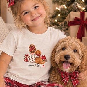 May include: A light heather gray short-sleeve t-shirt featuring a graphic of a child with a hair bow and a dog sitting by Christmas gifts. The text "Elonie & Rory" is printed below the graphic. The child wears a red plaid hair bow.