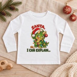K&ouml;nnte beinhalten: Ein wei&szlig;es Langarmshirt mit einer Grafik eines gr&uuml;nen Triceratops, der eine Weihnachtsmannm&uuml;tze tr&auml;gt und in bunte Lichterketten gewickelt ist. Der Dinosaurier steht neben einem Keks, mit dem roten Text SANTA, I CAN EXPLAIN... dar&uuml;ber und darunter.