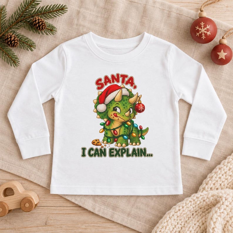 K&ouml;nnte beinhalten: Ein wei&szlig;es Langarmshirt mit einer Grafik eines gr&uuml;nen Triceratops, der eine Weihnachtsmannm&uuml;tze tr&auml;gt und in bunte Lichterketten gewickelt ist. Der Dinosaurier steht neben einem Keks, mit dem roten Text SANTA, I CAN EXPLAIN... dar&uuml;ber und darunter.