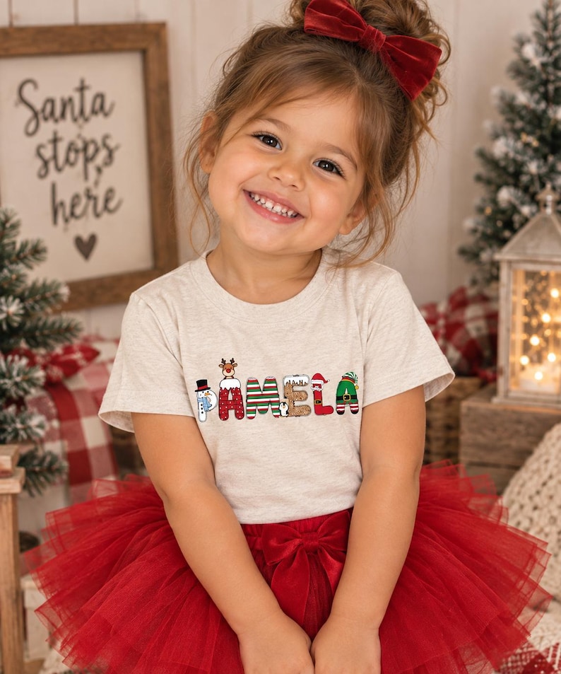 May include: A light heather gray short-sleeve t-shirt featuring the name PAMELA printed in a festive Christmas-themed font. Each letter is decorated with holiday motifs including a snowman, reindeer, candy stripes, and an elf hat.