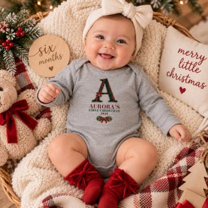 May include: A long-sleeved heather gray baby bodysuit featuring a large letter A in a green and red plaid pattern, with the text Aurora's First Christmas 2026 printed below. The bodysuit is paired with red socks featuring bows and a white headband.