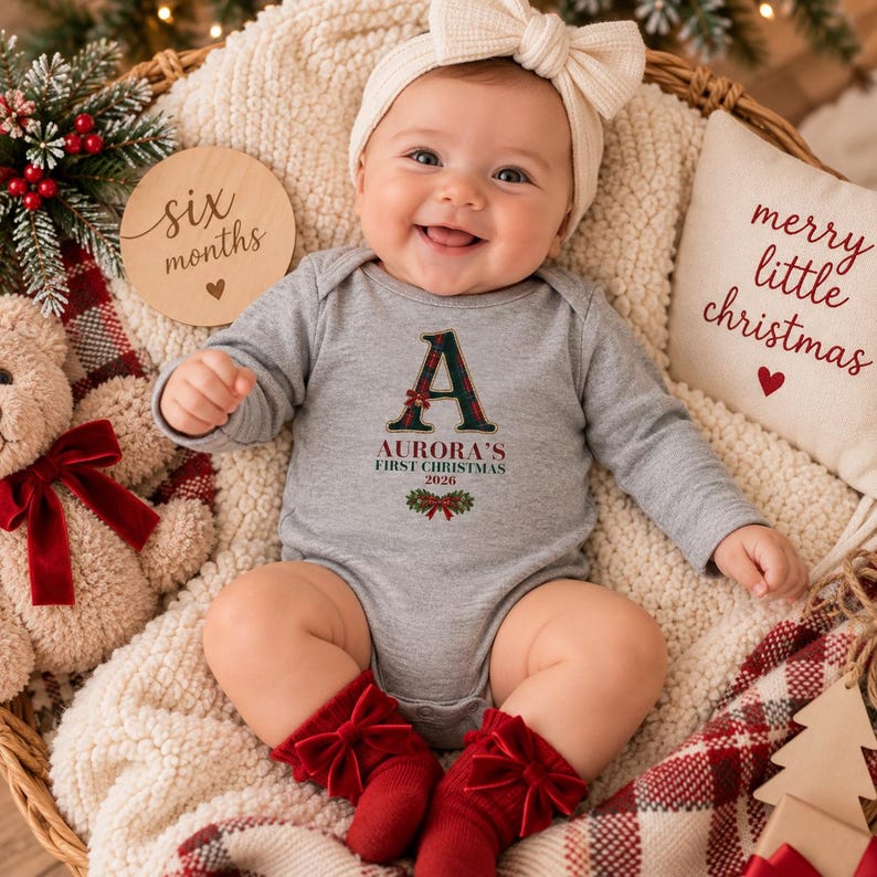 May include: A long-sleeved heather gray baby bodysuit featuring a large letter A in a green and red plaid pattern, with the text Aurora's First Christmas 2026 printed below. The bodysuit is paired with red socks featuring bows and a white headband.
