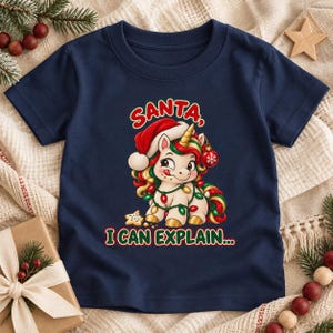 May include: A navy blue short-sleeve t-shirt featuring a graphic of a unicorn wearing a Santa hat and wrapped in Christmas lights. The unicorn has a gold horn and is holding a red ornament. The text reads SANTA, I CAN EXPLAIN...