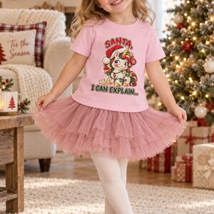 May include: A light pink short-sleeve t-shirt featuring a graphic of a unicorn wearing a Santa hat and wrapped in Christmas lights, with the text SANTA I CAN EXPLAIN. The shirt is paired with a layered, ruffled dusty rose tulle skirt.