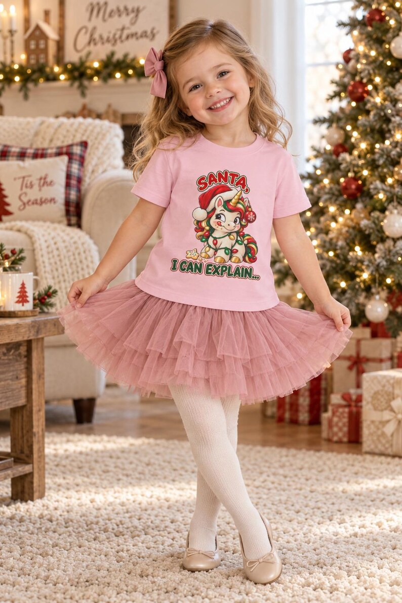 May include: A light pink short-sleeve t-shirt featuring a graphic of a unicorn wearing a Santa hat and wrapped in Christmas lights, with the text SANTA I CAN EXPLAIN. The shirt is paired with a layered, ruffled dusty rose tulle skirt.