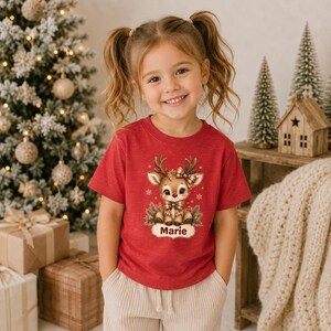 May include: A red short-sleeved t-shirt featuring a graphic of a cartoon reindeer with a plaid bow and scarf. The reindeer is surrounded by small snowflakes and pine branches. Below the reindeer, the name Marie is printed in a dark red font.