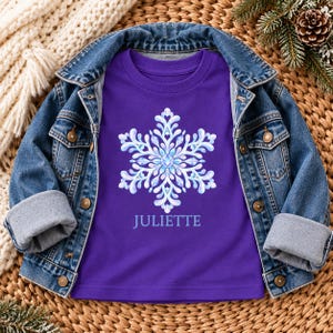 May include: A purple t-shirt featuring a large, intricate white and light blue snowflake graphic centered on the chest, with the name JULIETTE printed in white lettering directly below the snowflake design, layered under a blue denim jacket.