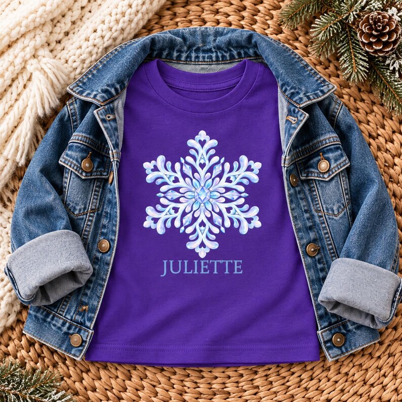 May include: A purple t-shirt featuring a large, intricate white and light blue snowflake graphic centered on the chest, with the name JULIETTE printed in white lettering directly below the snowflake design, layered under a blue denim jacket.