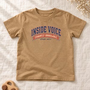May include: A tan short-sleeved t-shirt featuring the text INSIDE VOICE CURRENTLY UNAVAILABLE ETHAN AGE 3 in navy blue, white, and orange collegiate-style lettering and a banner graphic centered on the chest area of the garment.