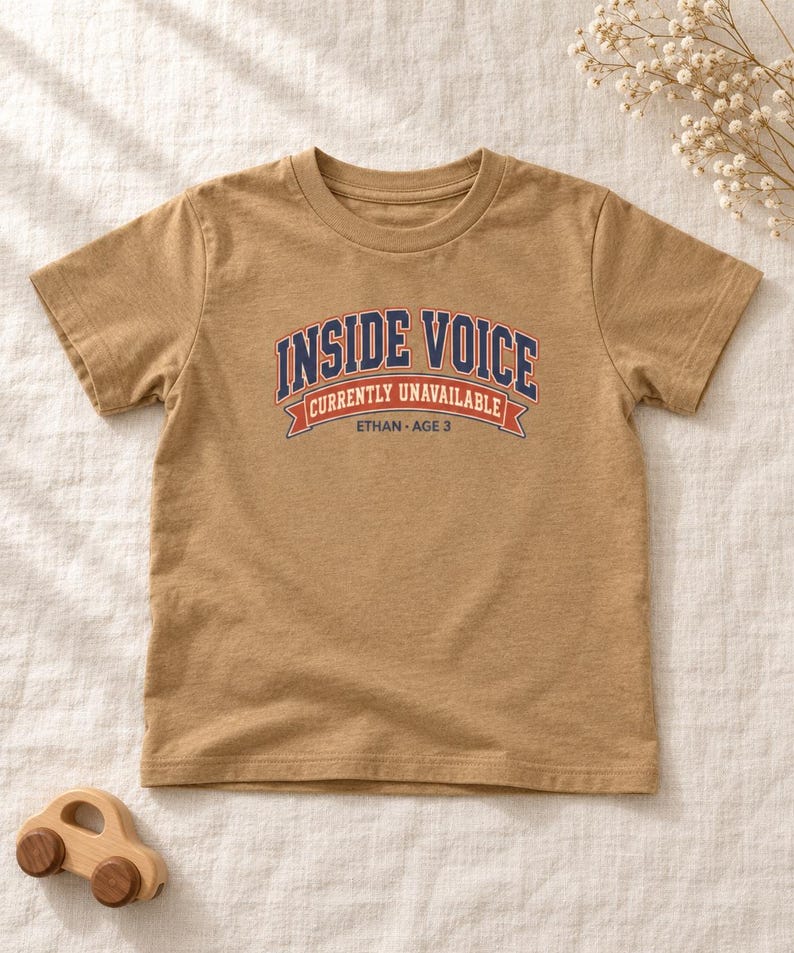 May include: A tan short-sleeved t-shirt featuring the text INSIDE VOICE CURRENTLY UNAVAILABLE ETHAN AGE 3 in navy blue, white, and orange collegiate-style lettering and a banner graphic centered on the chest area of the garment.