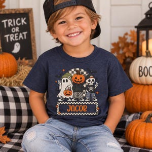 May include: A dark blue short-sleeve t-shirt featuring a graphic of a ghost, a pumpkin-headed character, and a skeleton. The design includes a checkered background, bats, and a rectangular label with the name JACOB in orange text.