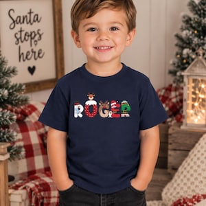 May include: A navy blue short-sleeved t-shirt featuring the name ROGER printed in large, decorative letters. Each letter incorporates festive holiday graphics, including a snowman, reindeer, Santa hat, and elf-themed patterns.