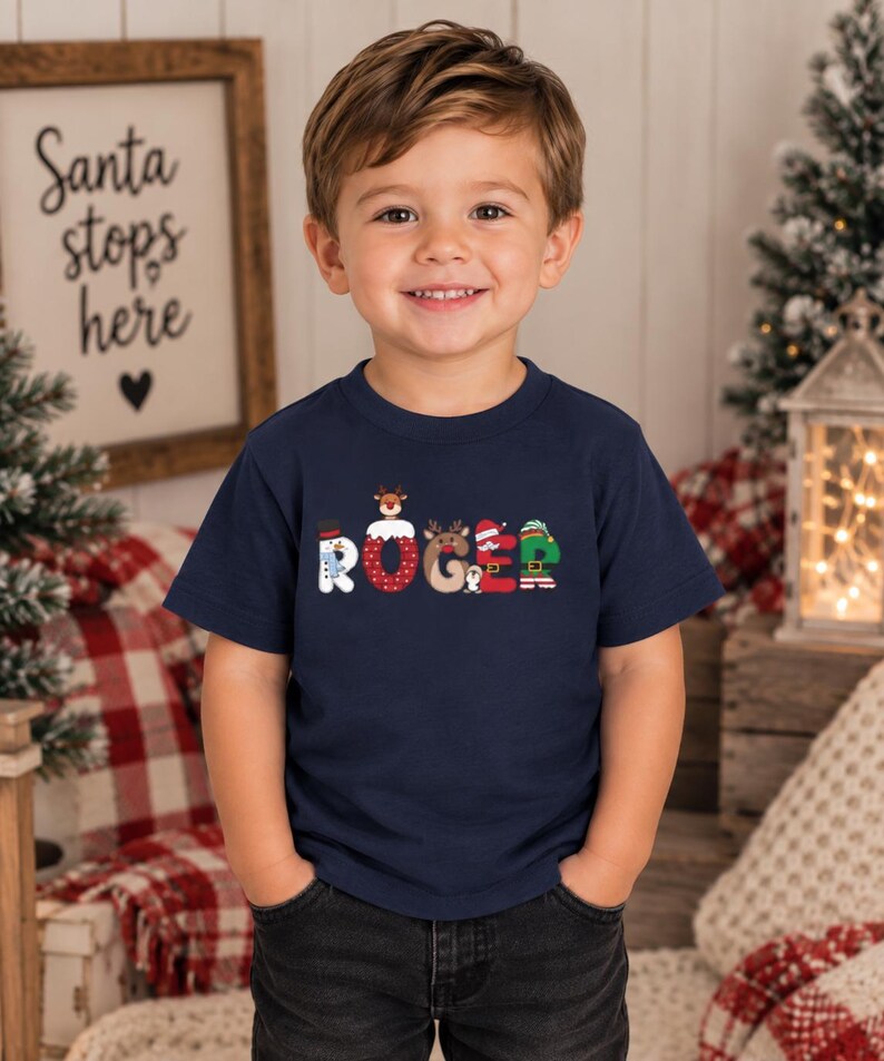 May include: A navy blue short-sleeved t-shirt featuring the name ROGER printed in large, decorative letters. Each letter incorporates festive holiday graphics, including a snowman, reindeer, Santa hat, and elf-themed patterns.