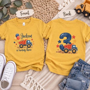 May include: Two mustard yellow t-shirts featuring construction truck graphics. The front displays a dump truck, balloons, and the text Jackson is turning three. The back shows a large number 3, a dump truck, and colorful stars.