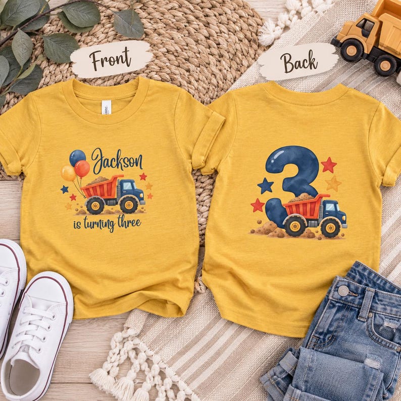 May include: Two mustard yellow t-shirts featuring construction truck graphics. The front displays a dump truck, balloons, and the text Jackson is turning three. The back shows a large number 3, a dump truck, and colorful stars.