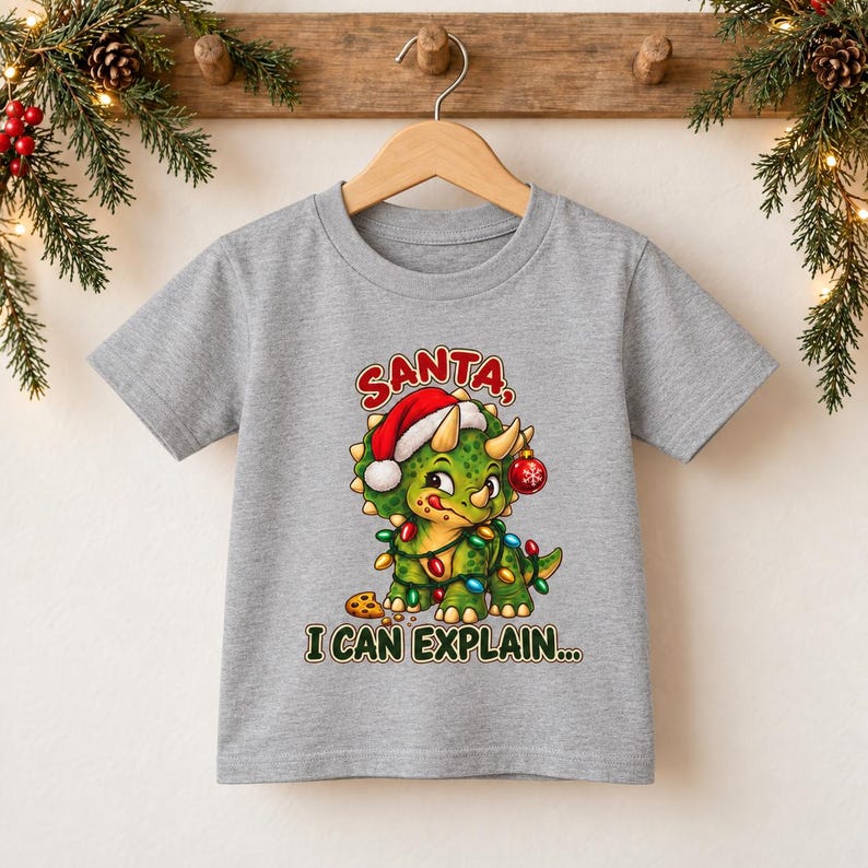 K&ouml;nnte beinhalten: Ein graues, meliertes Kurzarm-T-Shirt mit einem gr&uuml;nen Triceratops, der eine rot-wei&szlig;e Weihnachtsm&uuml;tze tr&auml;gt, in bunte Lichterketten verwickelt ist, eine rote Kugel h&auml;lt und neben einem Keks steht, mit dem Text SANTA, I CAN EXPLAIN...