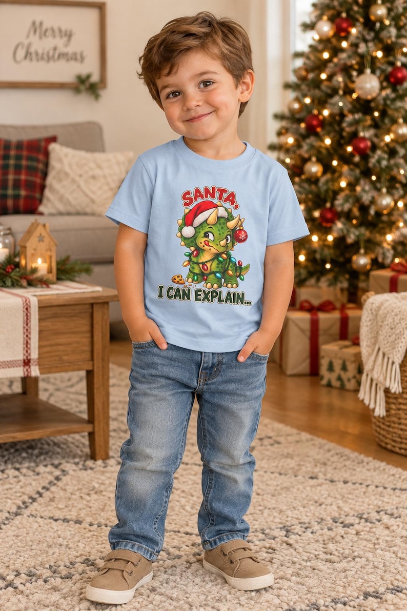 K&ouml;nnte beinhalten: Ein hellblaues T-Shirt mit kurzen &Auml;rmeln und einer Grafik eines gr&uuml;nen Triceratops, der eine rote Weihnachtsm&uuml;tze tr&auml;gt und mit bunten Lichterketten geschm&uuml;ckt ist. Der Text &uuml;ber dem Dinosaurier lautet SANTA und darunter I CAN EXPLAIN.