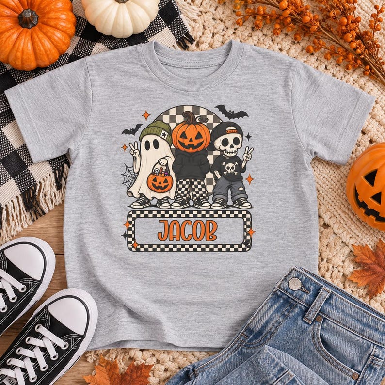 May include: A heather gray t-shirt featuring a graphic of a ghost, a pumpkin-headed figure, and a skeleton, all wearing hats and sneakers. The design includes bats, a spiderweb, and a rectangular box with the name JACOB in orange letters.