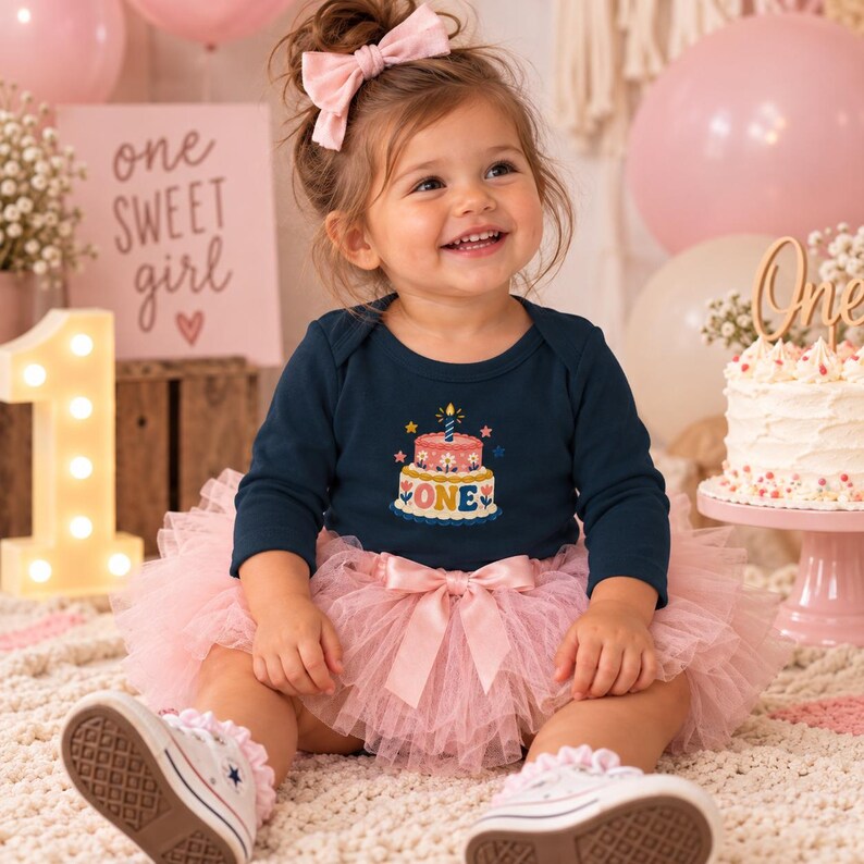 May include: A dark navy long-sleeve bodysuit featuring a graphic of a two-tiered birthday cake with a single candle and the word ONE, paired with a light pink layered tulle tutu skirt with a satin bow at the waist and white sneakers.