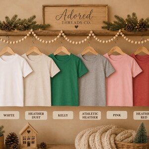 May include: Six short-sleeved crew neck t-shirts hanging on wooden hangers in a row. The colors from left to right are white, heather dust, kelly green, athletic heather gray, pink, and heather red. Each shirt is labeled with its color name.
