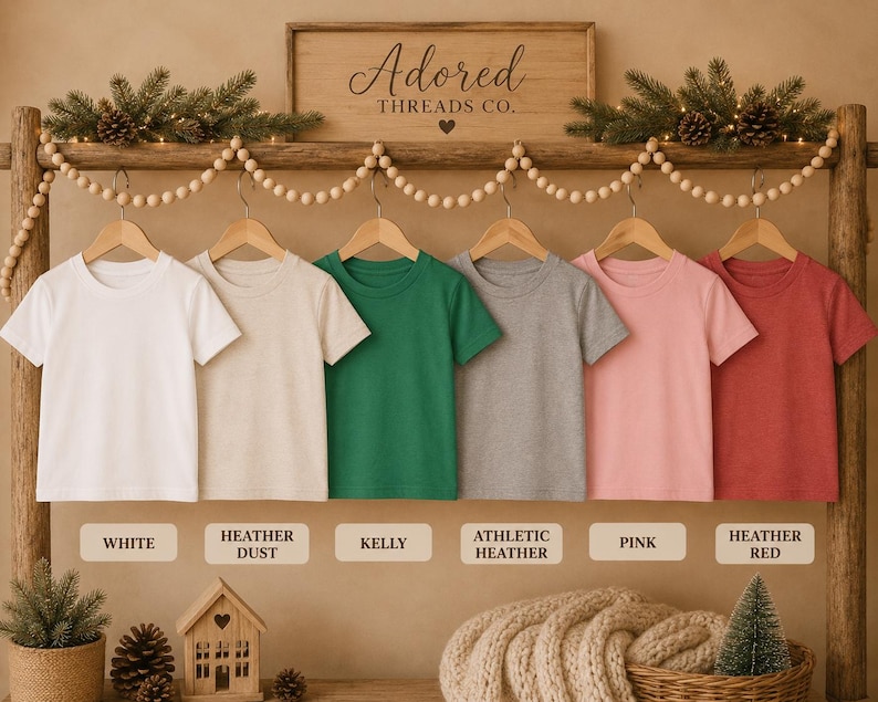 May include: Six short-sleeved crew neck t-shirts hanging on wooden hangers in a row. The colors from left to right are white, heather dust, kelly green, athletic heather gray, pink, and heather red. Each shirt is labeled with its color name.