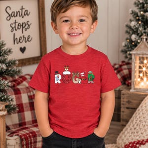 May include: A red short-sleeved t-shirt featuring the name ROGER printed in a festive holiday font. Each letter is stylized with Christmas-themed graphics, including a snowman, a reindeer, a Santa suit, and an elf outfit, all in vibrant colors.