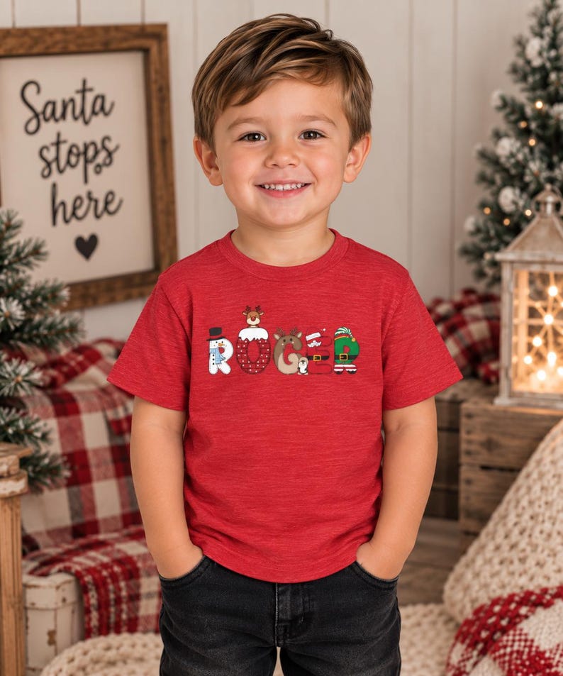 May include: A red short-sleeved t-shirt featuring the name ROGER printed in a festive holiday font. Each letter is stylized with Christmas-themed graphics, including a snowman, a reindeer, a Santa suit, and an elf outfit, all in vibrant colors.