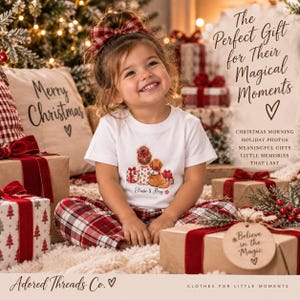 May include: A white short-sleeved t-shirt featuring a graphic of a person with a bun hairstyle sitting next to a golden dog, surrounded by Christmas gifts. The text reads 'Elenie & Rory' below the illustration, with a small red paw print.