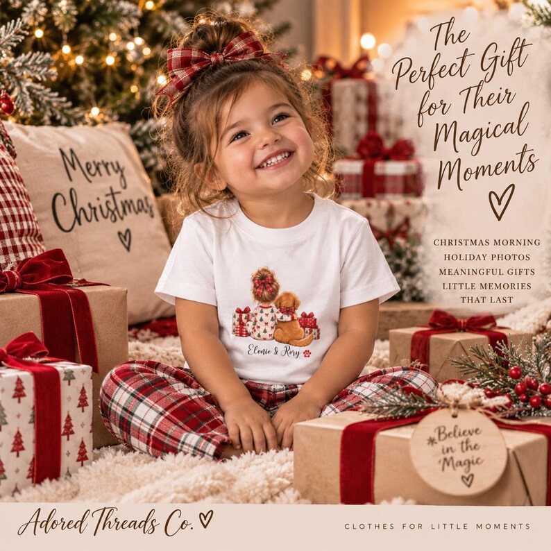 May include: A white short-sleeved t-shirt featuring a graphic of a person with a bun hairstyle sitting next to a golden dog, surrounded by Christmas gifts. The text reads 'Elenie & Rory' below the illustration, with a small red paw print.
