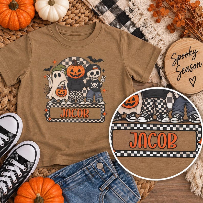 May include: A tan short-sleeve t-shirt featuring a graphic of a ghost, a pumpkin-headed figure, and a skeleton. The characters are wearing hats and sneakers. Below them is a checkered rectangular box with the orange text "JACOB" printed inside.